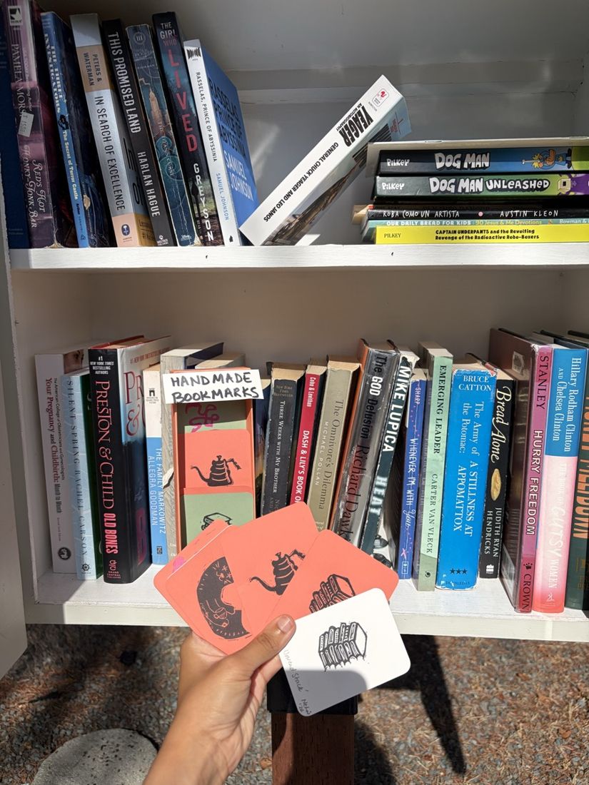 A hand fanning out little prints in front of a Little Free Library shelf, the handmade bookmarks dispenser tucked between the books behind