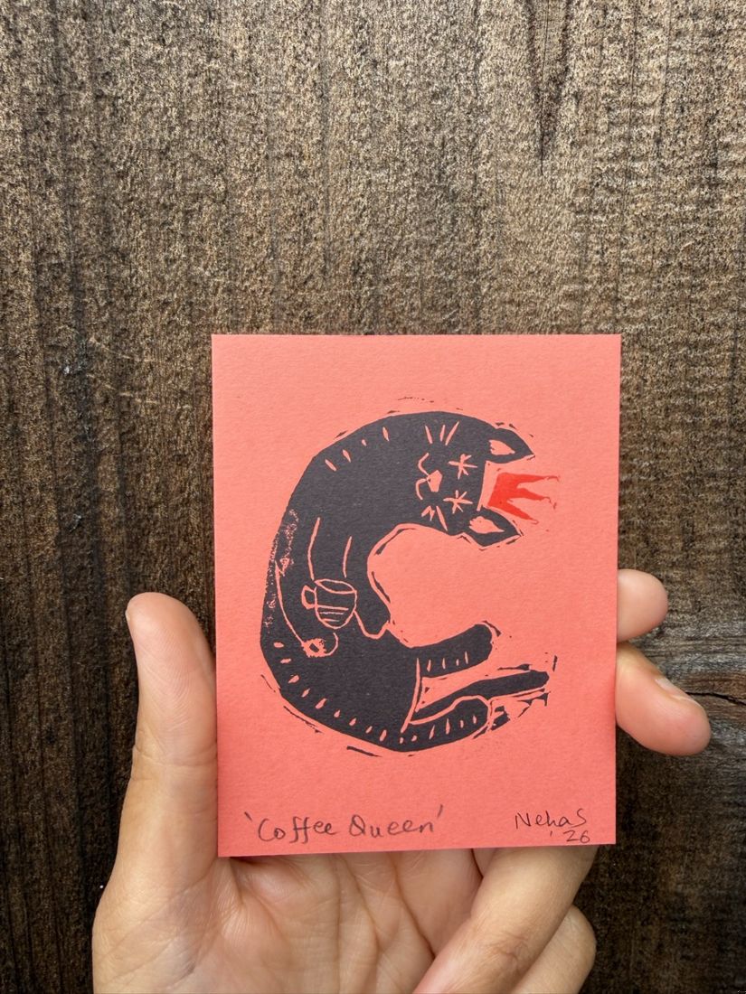 A tiny linocut print of a curled cat with a cup, titled Coffee Queen, on coral paper held in a hand