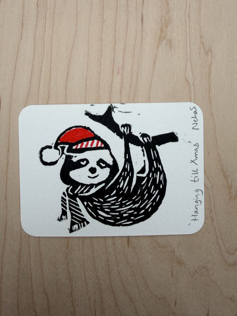 A tiny linocut print of a sloth in a Santa hat hanging from a branch, titled Hanging till Xmas