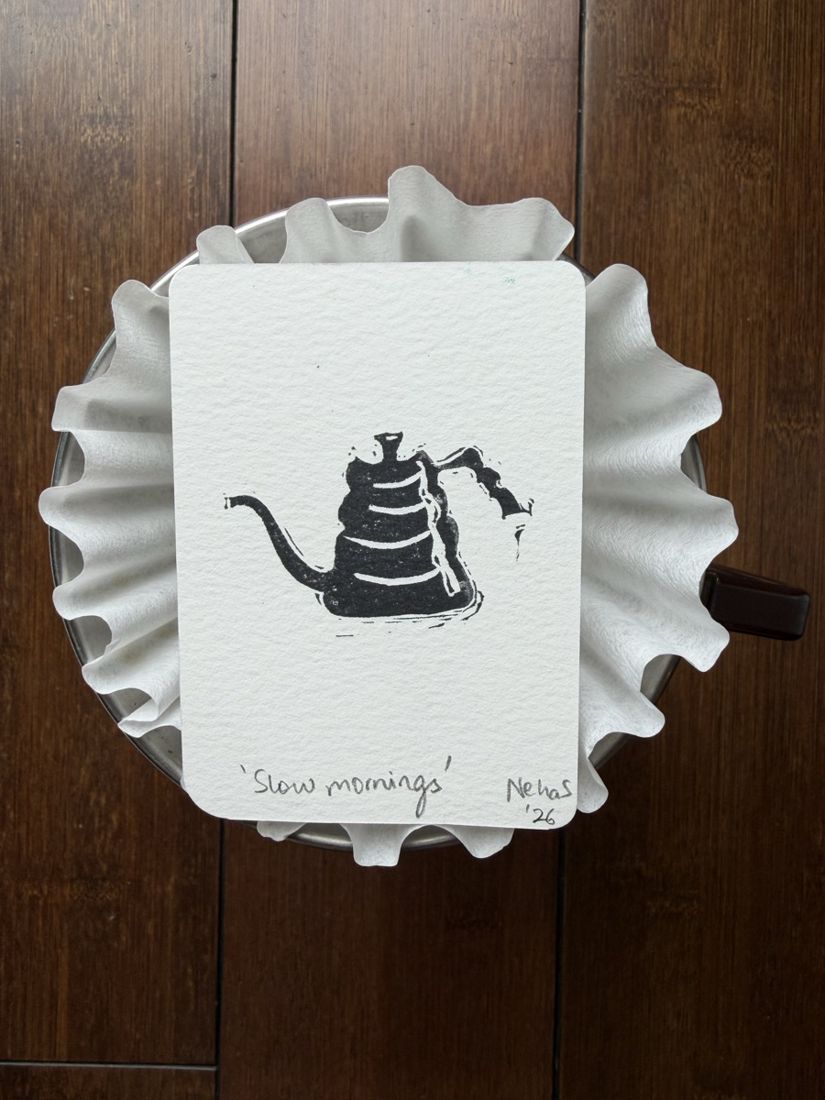 A tiny linocut print of a pour-over kettle titled Slow mornings, photographed on top of a coffee filter