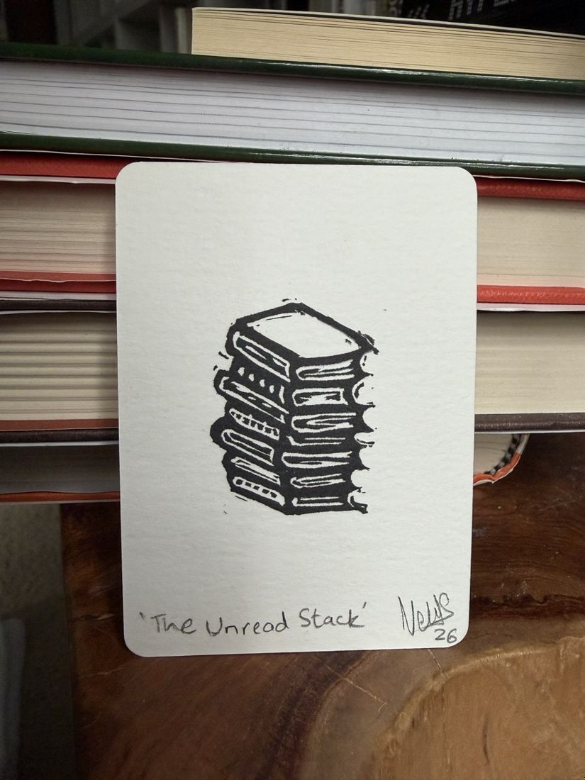 A tiny linocut print of a stack of books titled The Unread Stack, leaning against a shelf of real books