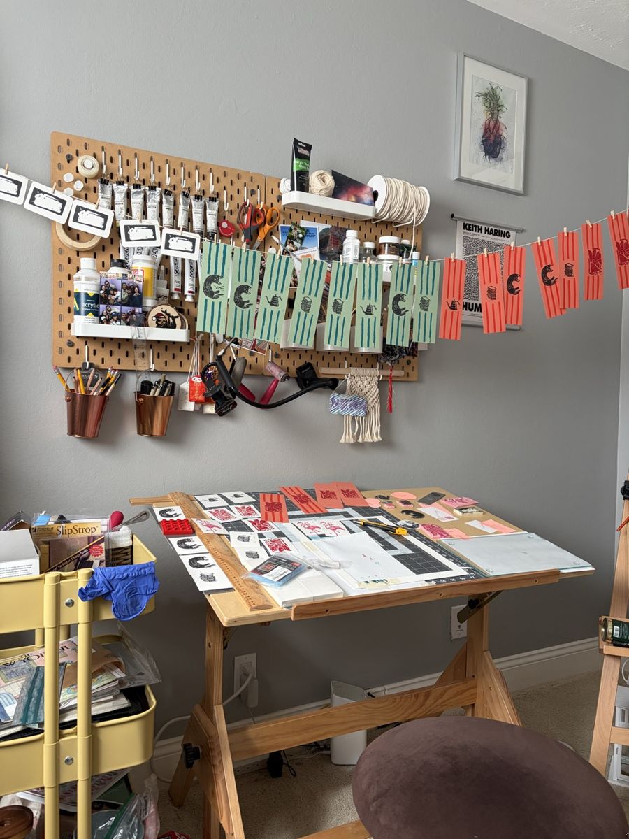 A corner of the home studio, bookmarks drying on a line strung across a pegboard of ink tubes and tools, a drafting table covered with prints in progress